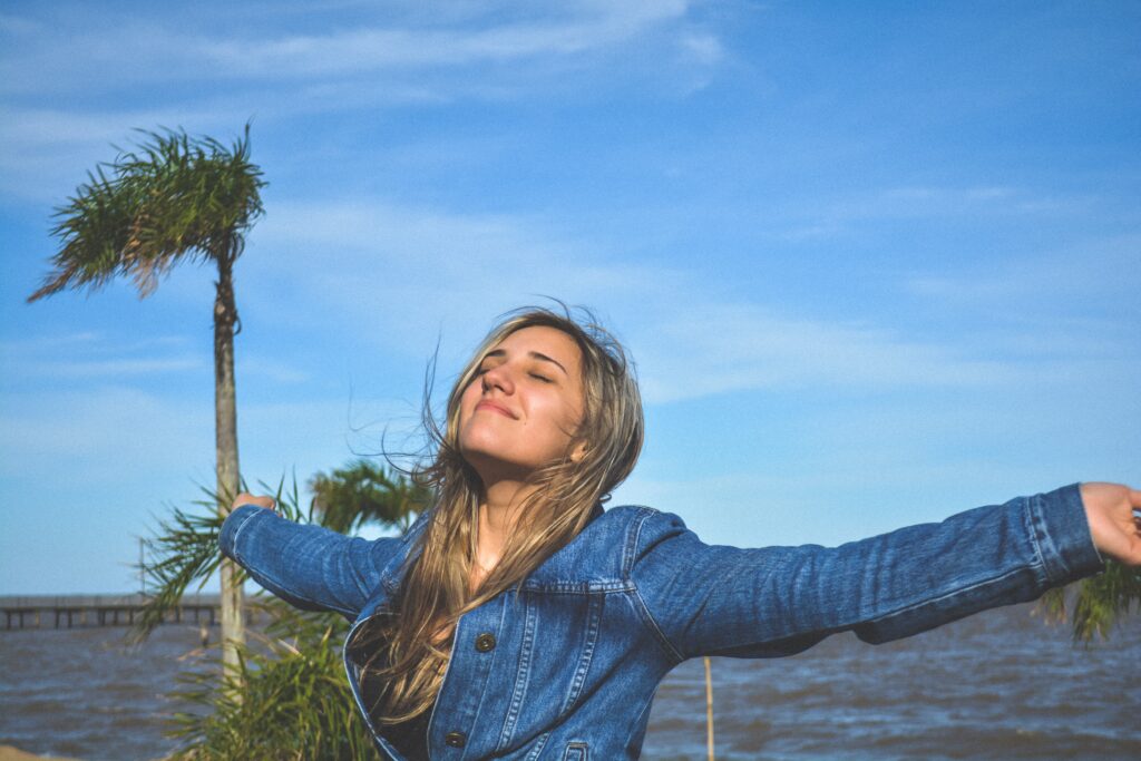 Young woman standing by the water's edge with her arms stretched out and a contented look on her face. 