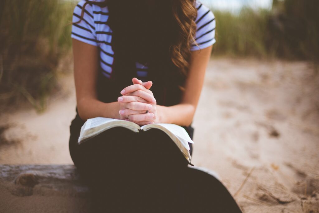 woman sitting on a log with hands folded over open Bible in her lap contemplating the words